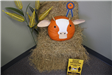 A pumpkin decorated as a cow sitting on a hay bale. Sign reads, Maintain social distancing of 1 cow