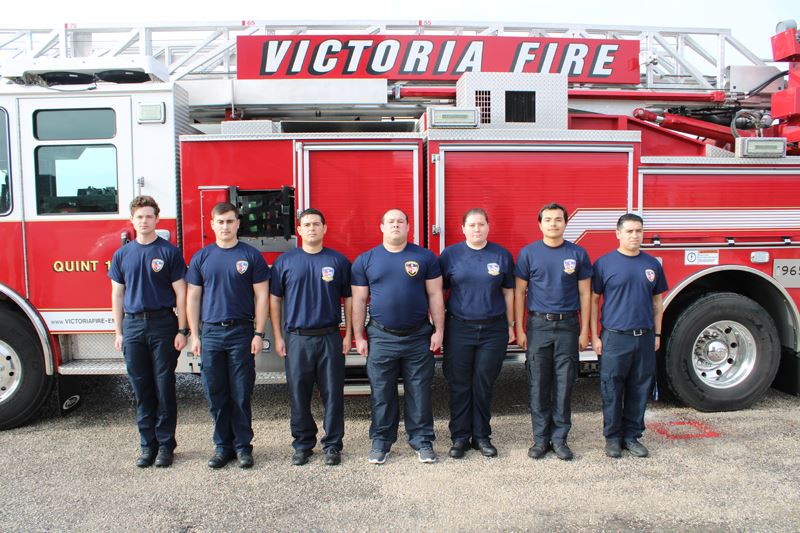 Seven people in fire department shirts stand in front of a fire truck