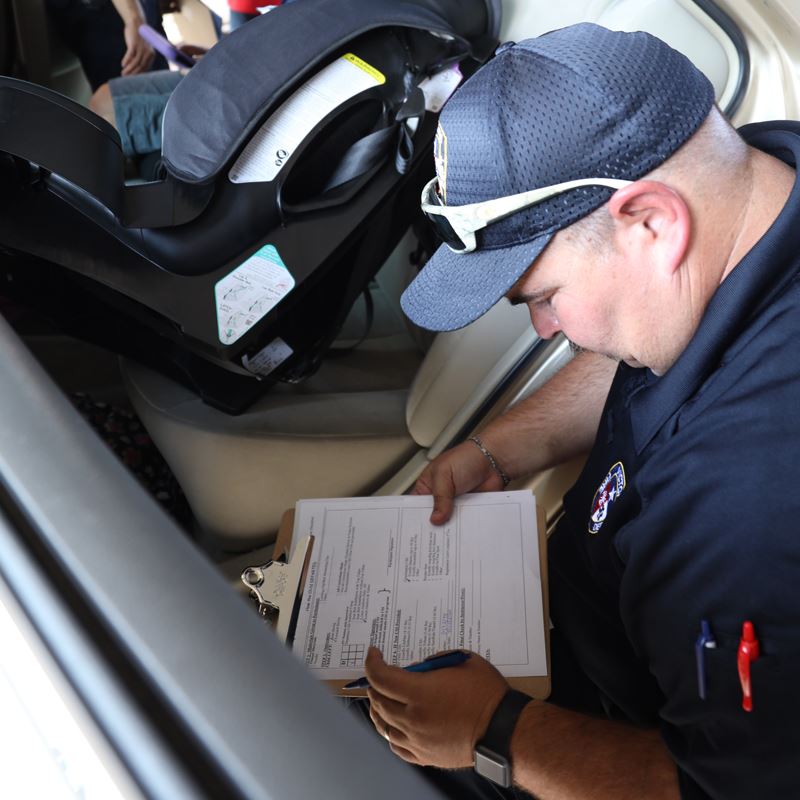 A man in a Victoria Fire Department polo crouches near a car seat and consults a checklist.