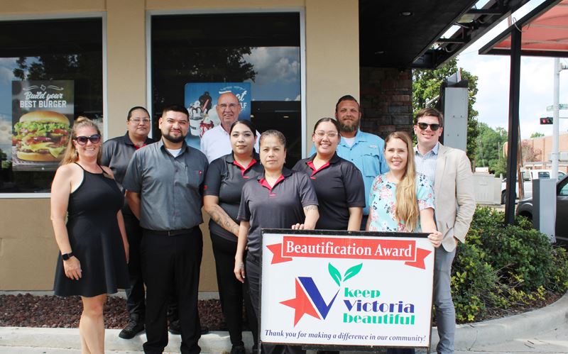 Group photo with KVB sign at Dairy Queen. Shrubbery and red volcanic rock bedding is visible.