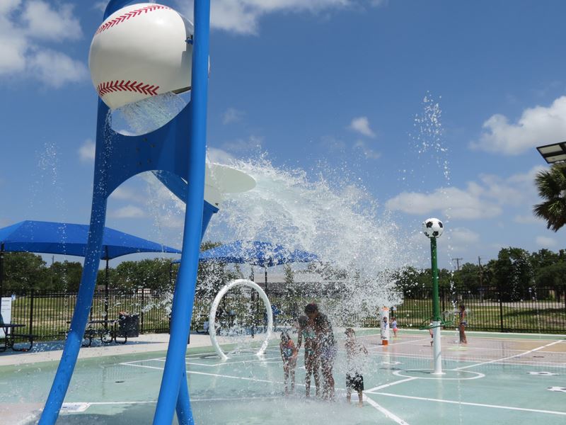 At a splash pad, a large bucket shaped like a baseball pours water on a group of kids