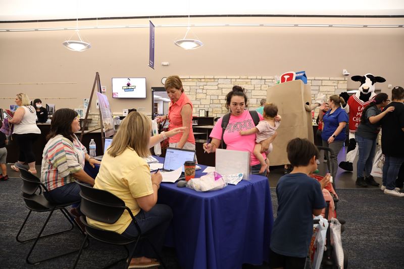 In a crowded library, two women, one of them holding a baby, approach a table booth setup.