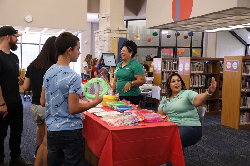 Two women in H-E-B polos at a booth in the library visiting with a group of people