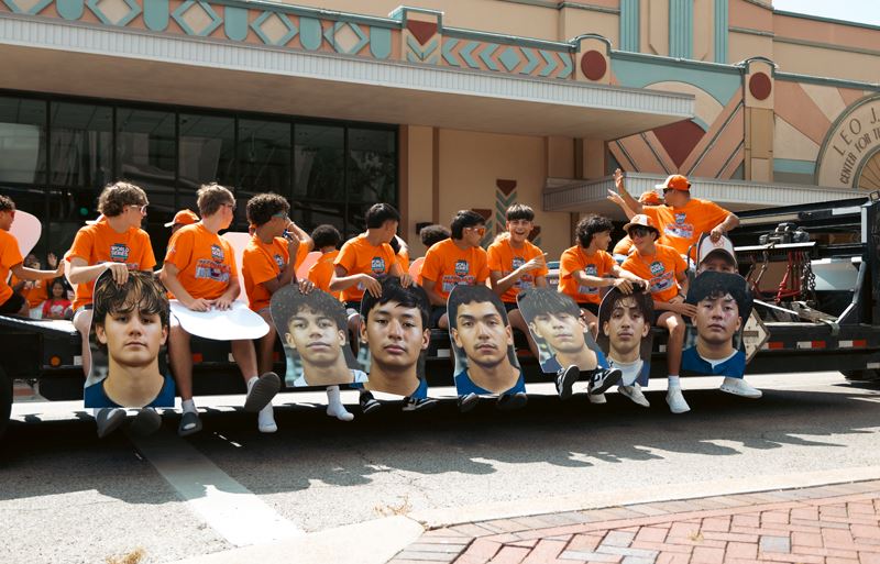 Teenage boys in matching orange shirts sit on a parade trailer holding cutouts of their faces