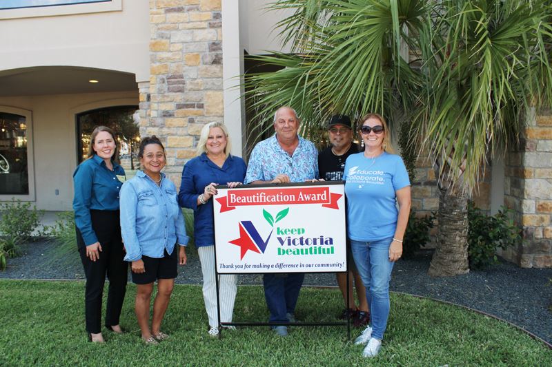 Group photo with KVB sign in front of a building with brick columns and a flowerbed with a palm tree