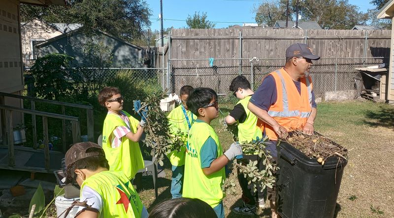 A man and five kids in work vests collect small branches and put them in a trash can