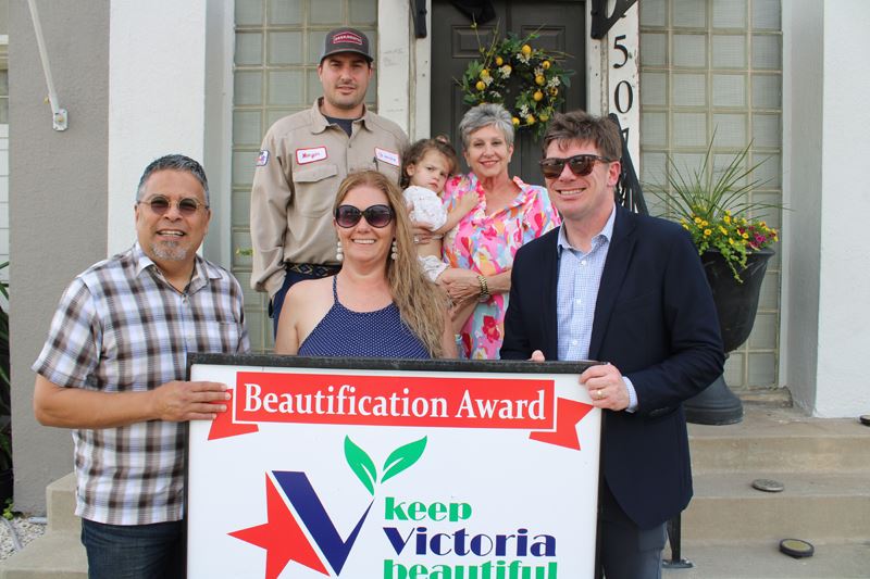 Group of people holding a KVB sign on a front porch with a decorative flower pot and wreath