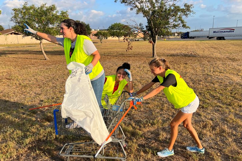 A young woman pushing another young woman in a shopping cart while another young woman salutes