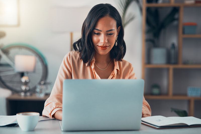 Woman using a laptop in a home office setting with a cup of coffee and an open notebook