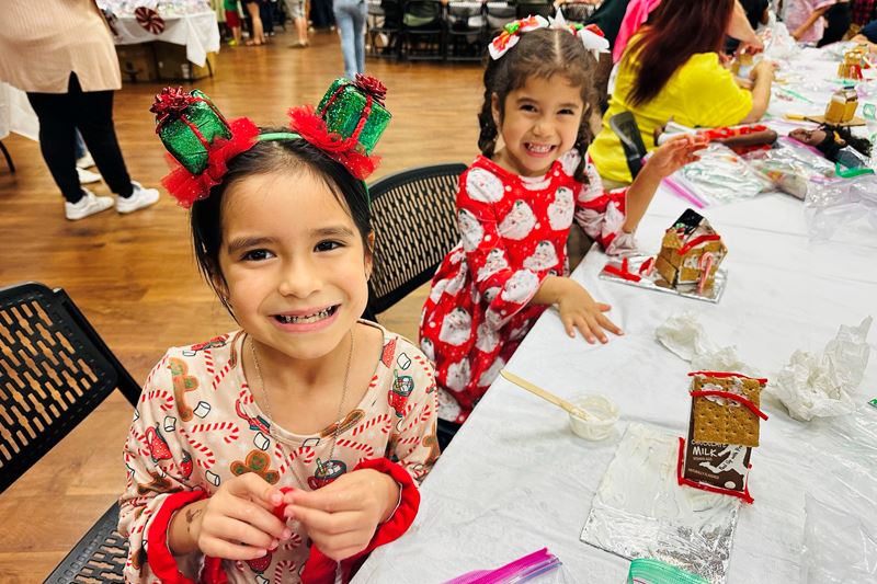 Two young girls in Christmas dresses smile while building gingerbread houses