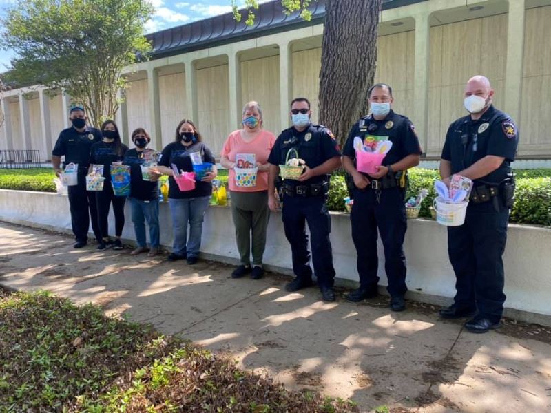 A group including uniformed police officers stands along an outdoor wall holding Easter baskets