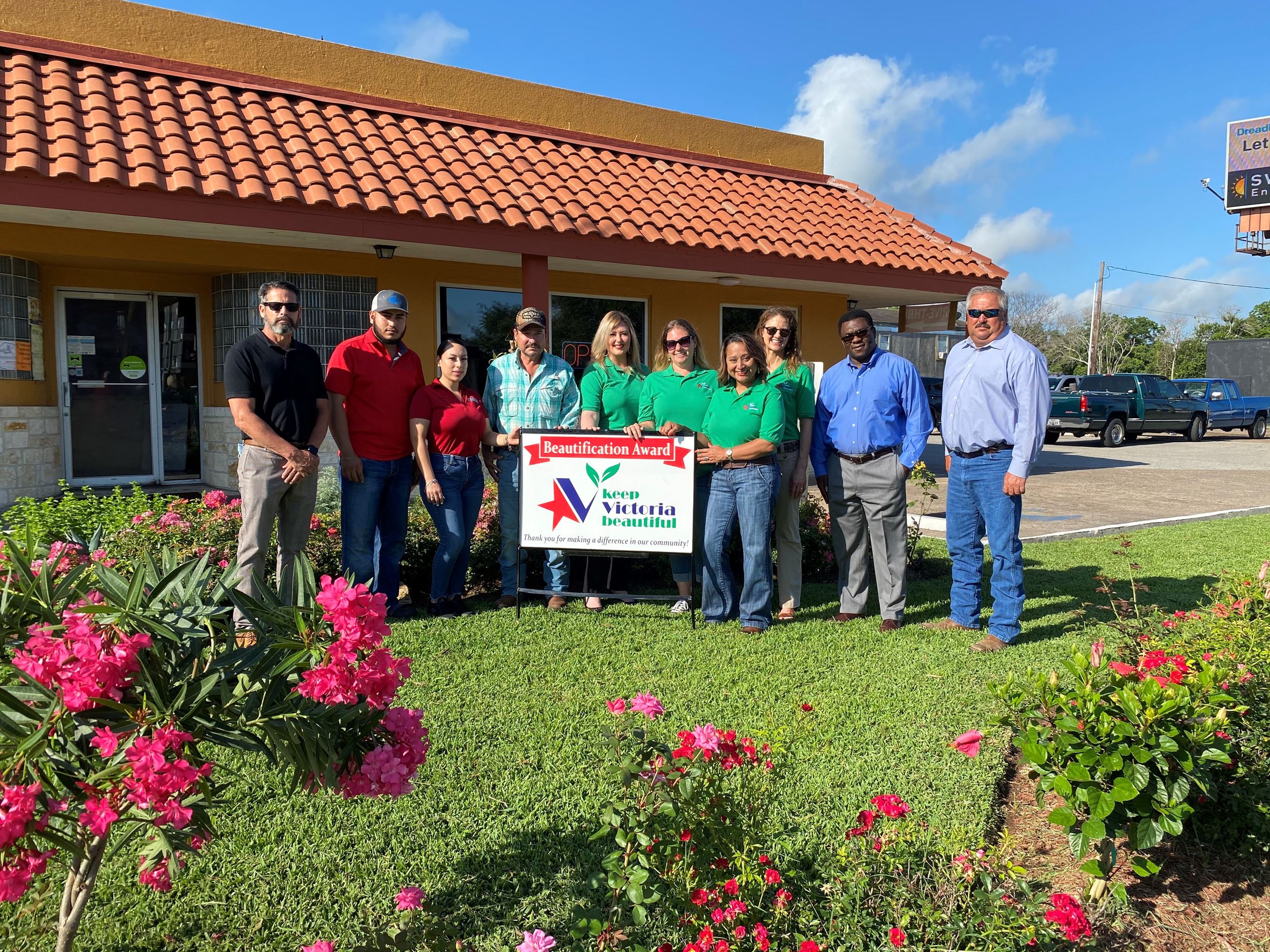 Ten people pose with a sign that says Business Beautification Award. Flowering plants in foreground.