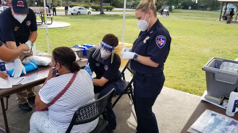 Fire department employee gives a woman a flu shot in an outdoor park. Other fire employees are near.