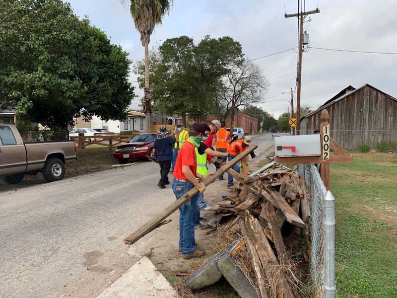 People in safety vests pile large pieces of wood along a fence by the street.