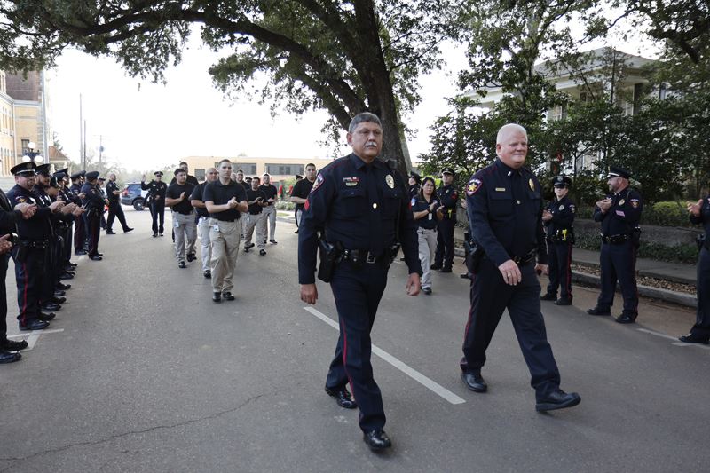 Two police officers walk down the street, flanked by rows of police officers applauding them.