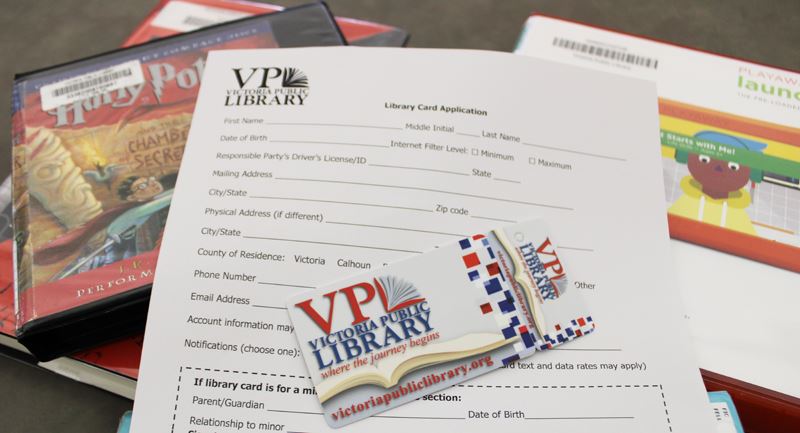 A library card and application form sit on a pile of books, an audiobook and a tablet.