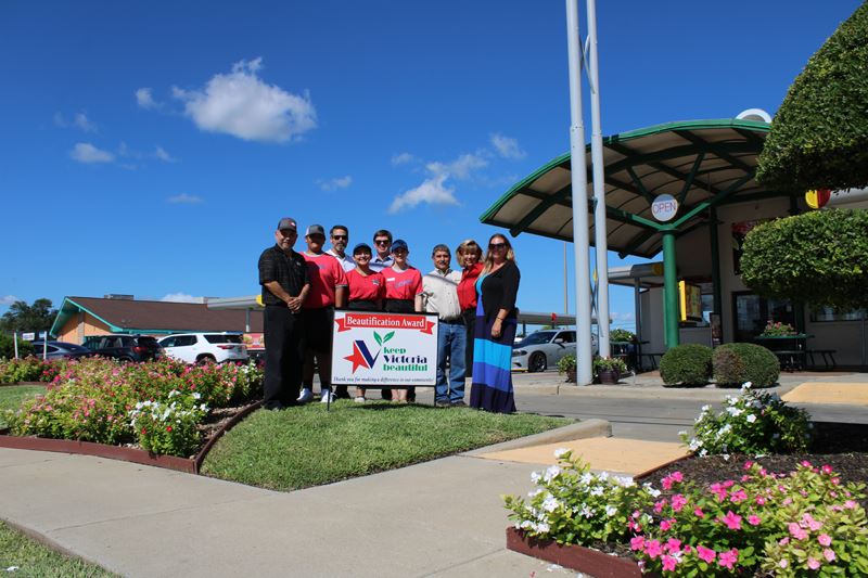 Group photo with KVB sign at Sonic on Navarro. Landscaping includes flowerbeds and trimmed bushes.