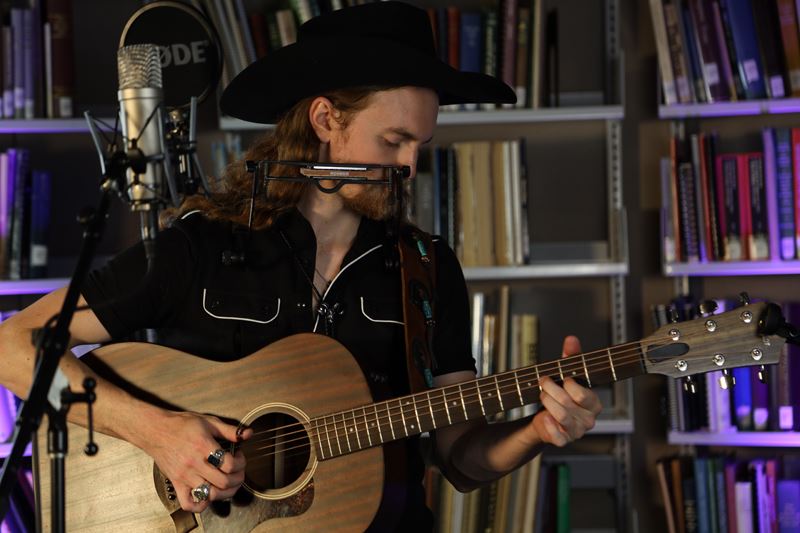 Man with a black cowboy hat plays a guitar. He also wears a harmonica holder to play harmonica.