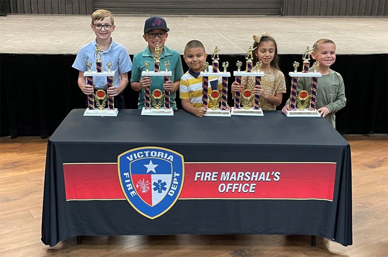 Five kids stand at a table with trophies. The table has a tablecloth that says Fire Marshal's Off