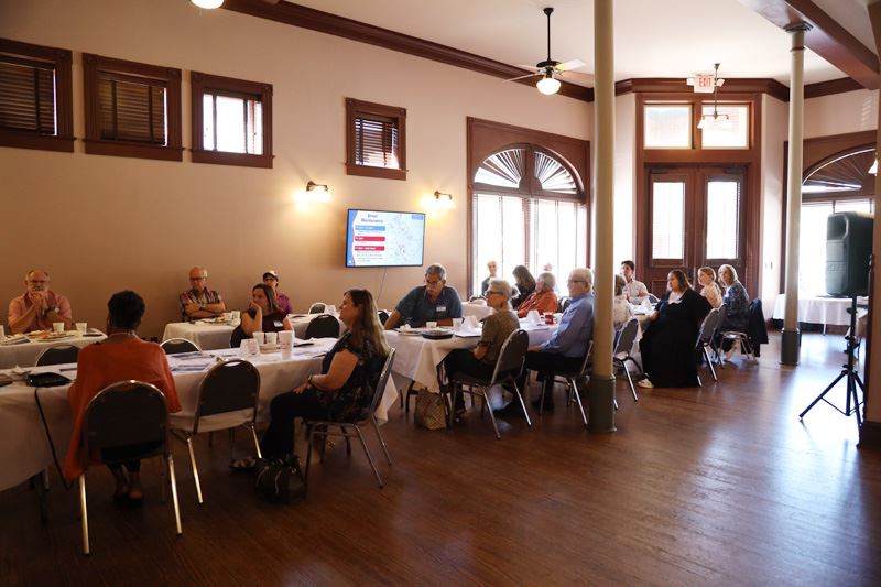 People sitting at rectangle tables look toward the front of the room.