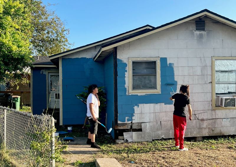 Two teenagers use paint rollers to paint a white house blue