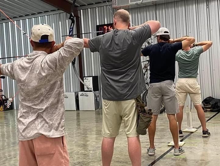 Four archers in a large indoor area line up and take aim