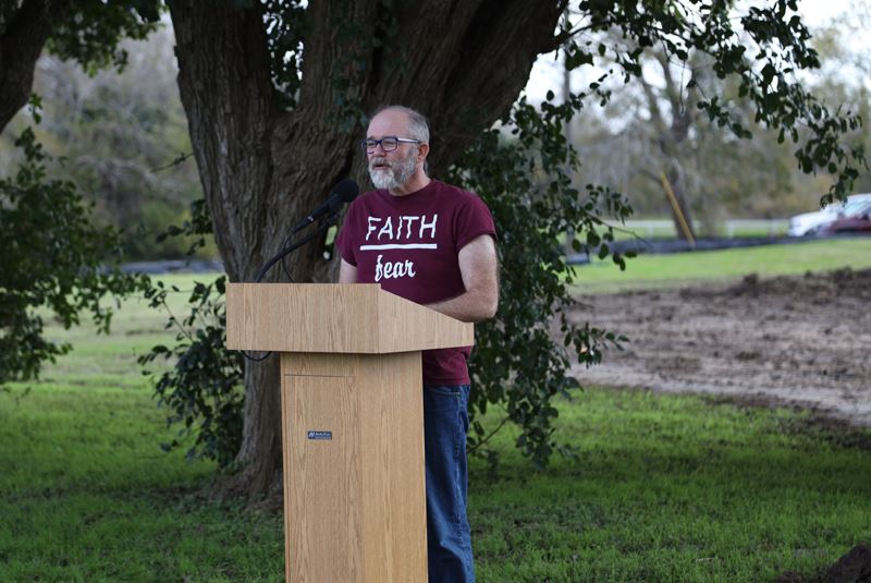 A man stands in front of a podium in a park with a tree and overturned dirt behind him Opens in new window