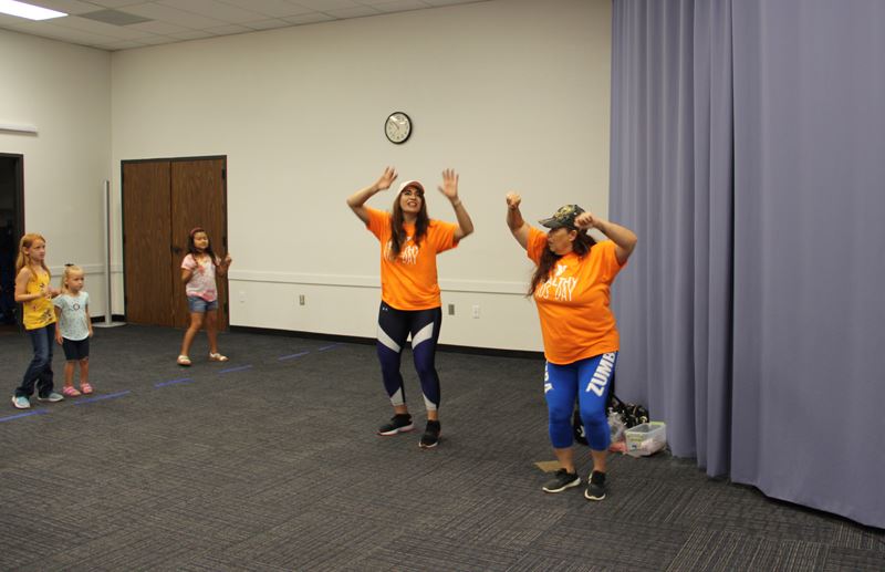 Two women wearing workout pants and orange T shirts lead kids through dancing motions.