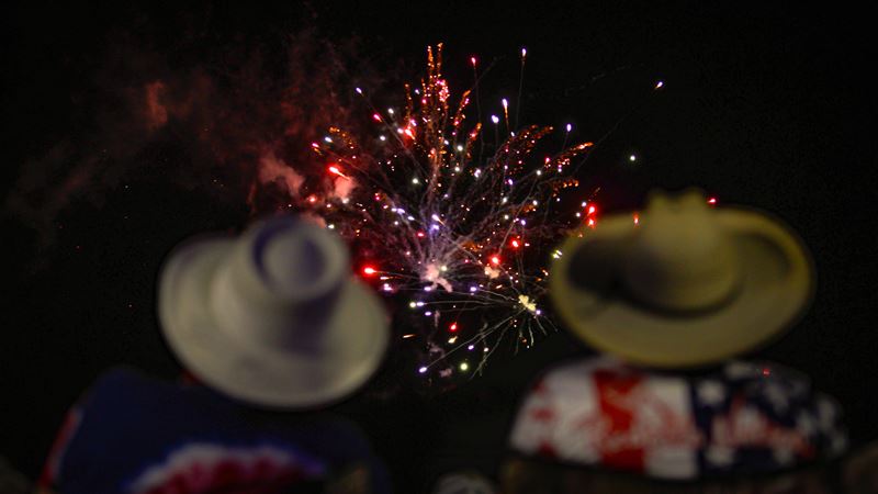 Two people wearing red, white and blue shirts and wide-brimmed hats watch a fireworks show Opens in new window