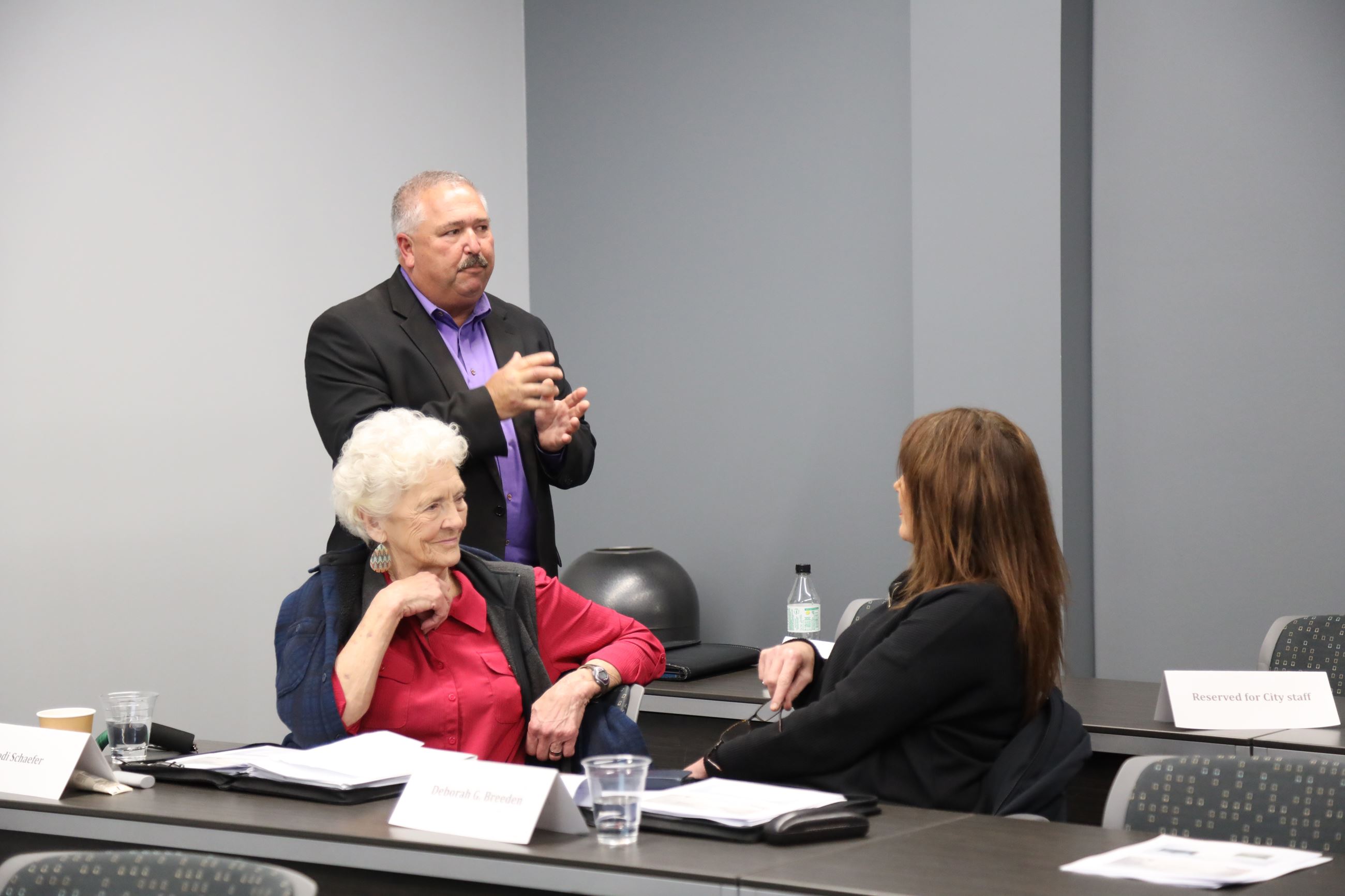 A man standing speaks with two women seated at a table