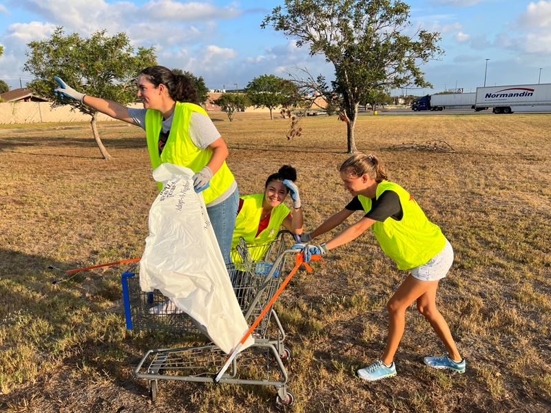 A young woman pushing two other young women in a shopping cart. One sitting, one standing
