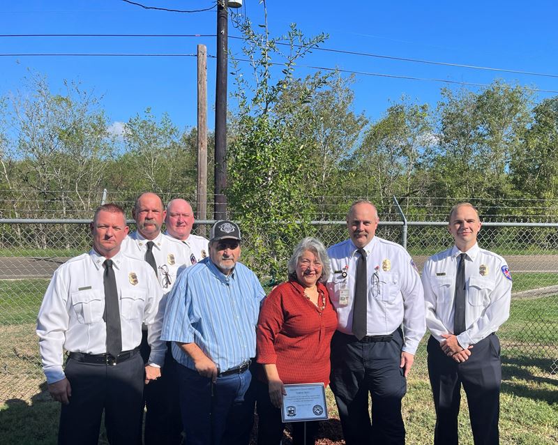 Group photo in front of a young tree. Five in fire department dress clothes, two in casual clothes Opens in new window