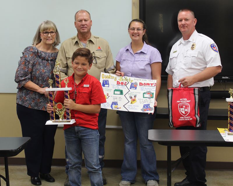 A boy holding a trophy. Four adults stand behind him holding a poster and a red bag.