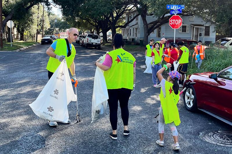 A group of children and adults walk through neighborhood holding trash bags and wearing safety vests Opens in new window