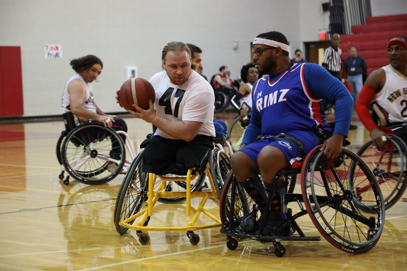 People playing wheelchair basketball. A man in a white jersey defends from a man in a blue jersey.
