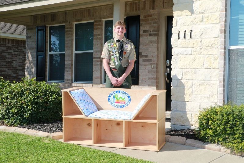 Teen in Boy Scout uniform poses with a wooden reading nook that features bookshelves.
