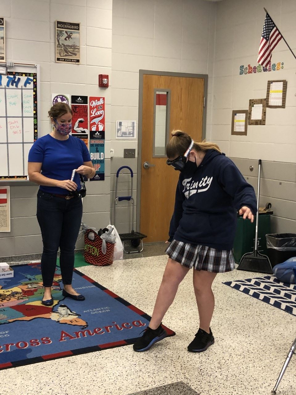 A woman watches a uniformed girl wearing impairment goggles try to walk in a line in a classroom.