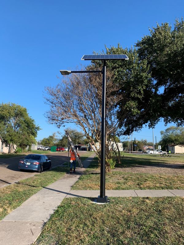 A streetlight by a sidewalk is topped with a wide solar panel. A worker approaches with a ladder.