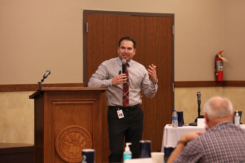 Man with a tie leans on a podium and speaks into a handheld microphone